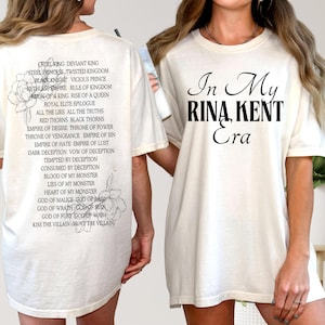 May include: Cream-colored t-shirt with black text that reads "In My Rina Kent Era" on the front. The back of the shirt features a list of phrases and flower illustrations. A casual, comfortable tee.