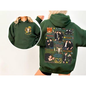 May include: A dark green hoodie with a front pocket and a hood. The front features a graphic of a skull and text. The back has multiple illustrations and text, including a bear, motorcycle, and couples. The text includes phrases like "I love you" and "YA S TOBOY."
