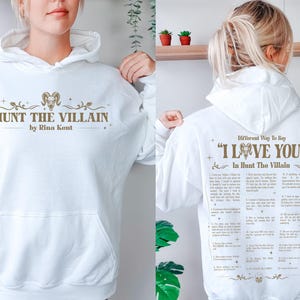 May include: A white hooded sweatshirt with the text "HUNT THE VILLAIN" and "by Rina Kent" in a gold color on the front. The back of the sweatshirt has the text "Different Way To Say 'I LOVE YOU' In Hunt The Villain".