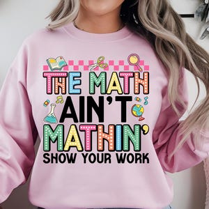 May include: A light pink sweatshirt with the text "THE MATH AIN'T MATHIN' SHOW YOUR WORK" in colorful, playful lettering. The design includes school-themed graphics like a book, scissors, and a magnifying glass.