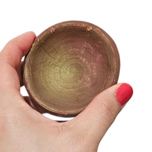 Puede incluir: Un cuenco pequeño y redondo hecho a mano con una superficie texturizada. El cuenco se sostiene en una mano y tiene un exterior marrón rojizo y un interior amarillo dorado. El borde del cuenco tiene una textura ligeramente irregular.