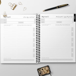 May include: A white planner with a black spiral binding. The planner has two pages open, each with the title "Big Goal" and space for writing down goals, milestones, and action plans. The text "Dream big, work hard" is written on the left page and "Think positive, enjoy the journey" is written on the right page.