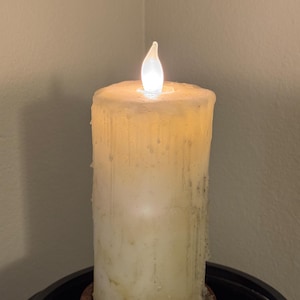 May include: A cylindrical off-white pillar candle with a textured, dripped wax appearance. The candle features a small, glowing LED flame at the top, emitting a soft light that illuminates the upper portion of the wax body.