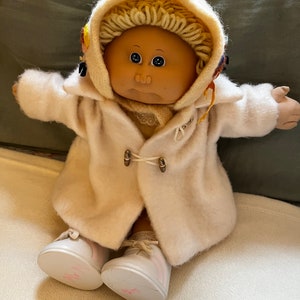 May include: A Cabbage Patch Kid doll wearing a white coat with a hood, a white scarf, and white shoes. The doll has blonde hair and brown eyes.
