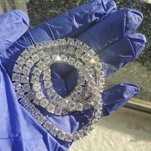 Op de afbeelding: Een sprankelende, zilverkleurige tennis ketting met talrijke ronde, heldere edelstenen. De ketting wordt gepresenteerd tegen een blauwe achtergrond, die de schittering benadrukt. De edelstenen zijn dicht op elkaar geplaatst, waardoor een doorlopende lijn ontstaat.