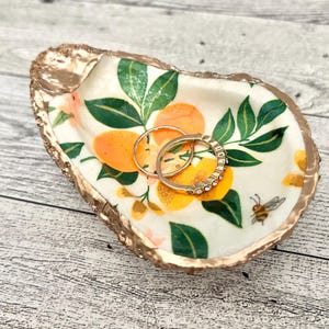 May include: A white ceramic oyster shell dish with a gold rim and a citrus fruit and bee design. Two gold rings are sitting in the dish.