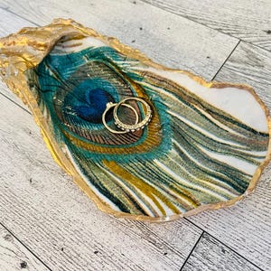 May include: A gold-rimmed oyster shell dish with a peacock feather design. Two gold rings are resting in the dish.