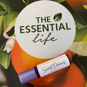 Könnte beinhalten: Eine violette Rollflasche mit einem weißen Etikett, auf dem "Sweet Dreams Essential Oil 100% Natural Product CPTG" steht. Die Flasche liegt auf einem Hintergrund aus orangefarbenen und grünen Blättern.