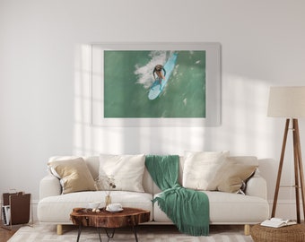 May include: A white framed photograph of a surfer riding a wave. The photo is hanging on a white wall above a white couch with a green throw blanket.