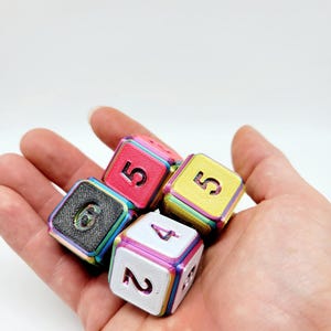 May include: A set of four multi-colored dice with rainbow-toned edges and textured faces. The dice feature black, pink, yellow, and white surfaces with embossed numbers including 6, 5, 5, 4, and 2 clearly visible on the top and side faces.
