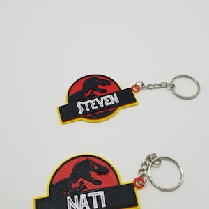 May include: Two keychains featuring a dinosaur silhouette within a red circle, with the names "STEVEN" and "NATI" on black rectangles. The keychains have yellow borders and silver key rings.