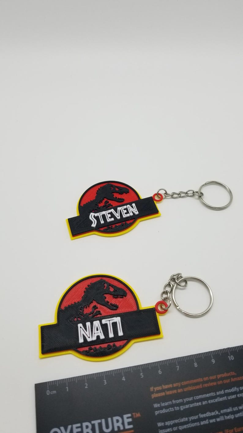 May include: Two keychains featuring a dinosaur silhouette within a red circle, with the names "STEVEN" and "NATI" on black rectangles. The keychains have yellow borders and silver key rings.