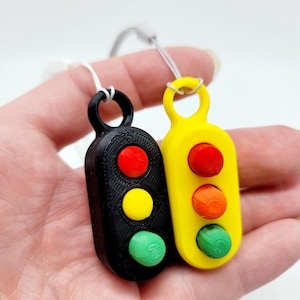 May include: Two miniature traffic light pendants, one black and one yellow, each with three colored lights: red, yellow, and green. The pendants are held by a person's hand and are attached to white cords.