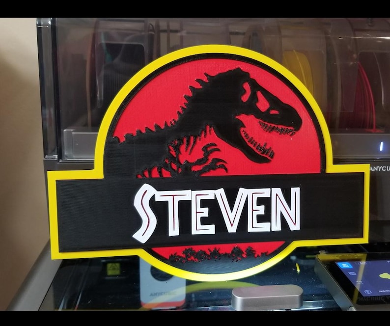 May include: A 3D-printed sign featuring a black Tyrannosaurus Rex silhouette on a red background, framed by a yellow border. The name "STEVEN" is displayed in white, block letters on a black rectangular base.