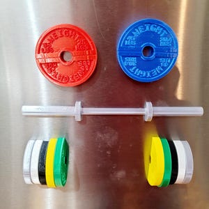 May include: A set of barbell-themed magnets featuring a red weight plate labeled 55 LBS 25 KGS, a blue plate labeled 45 LBS 20.4 KGS, a white barbell bar, and two stacks of colorful weight plates in yellow, green, black, white, and silver.