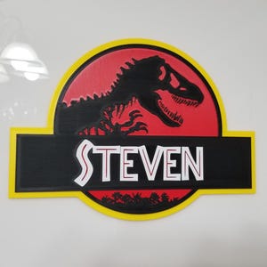 May include: A wooden sign featuring the Jurassic Park logo with a black Tyrannosaurus Rex silhouette on a red background. The name "STEVEN" is displayed in white letters on a black banner, framed by a yellow border.