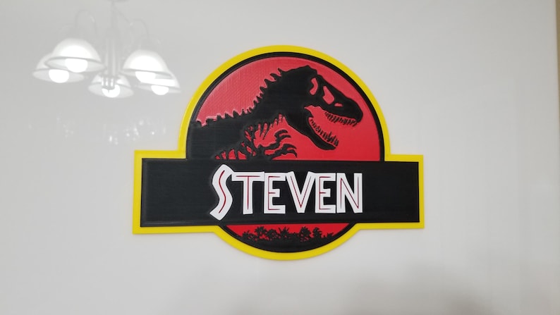 May include: A wooden sign featuring the Jurassic Park logo with a black Tyrannosaurus Rex silhouette on a red background. The name "STEVEN" is displayed in white letters on a black banner, framed by a yellow border.
