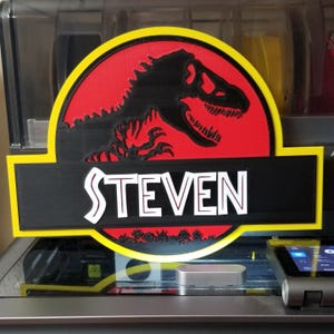 May include: A custom sign featuring the Jurassic Park logo with a black Tyrannosaurus Rex silhouette on a red background. The name "STEVEN" is displayed in white block letters on a black banner, framed by a yellow border.