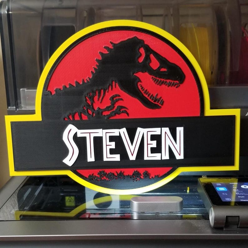 May include: A custom sign featuring the Jurassic Park logo with a black Tyrannosaurus Rex silhouette on a red background. The name "STEVEN" is displayed in white block letters on a black banner, framed by a yellow border.