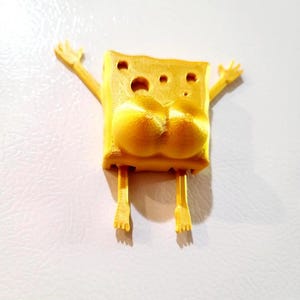 May include: A yellow 3D-printed figurine of a cartoon sponge character with arms raised and legs extended. The character has a porous body, and the design includes exaggerated features.