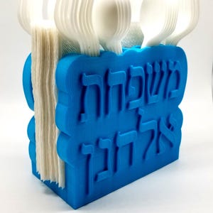 May include: A blue 3D-printed utensil holder with Hebrew text. The holder contains white plastic forks, spoons, and a stack of napkins. The design features a wavy top and bottom edge.