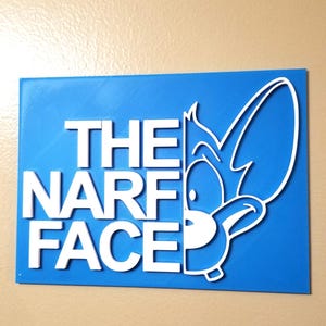 Op de afbeelding: Een blauw rechthoekig bord met de witte woorden "THE NARF FACE". Aan de rechterkant van het bord staat een wit stripfiguur met een groot oor en een lachend gezicht.