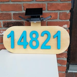 May include: Wooden address sign with the number 14821 in bright blue against a light wood oval. The sign is mounted on a white surface in front of a brick wall and a dark wooden door. A black solar panel is mounted above the sign.