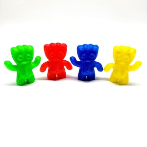 May include: Four colorful gummy candy figures in green, red, blue, and yellow. Each figure has a smiling face, arms outstretched, and a textured head. The candies are arranged in a row against a white background.