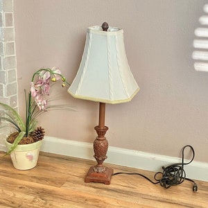 May include: A wooden table lamp with a white fabric shade. The lamp is on a wooden floor with a rug underneath. There is a potted plant with pink flowers in the background.