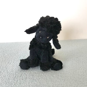 May include: A black plush toy poodle sitting on a light blue fuzzy surface. The poodle has a curly black coat and black eyes.