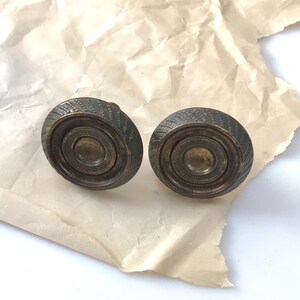 May include: Two antique brass door knobs with a detailed, textured design. The knobs have a circular shape with concentric rings and a patterned outer edge. They are set against a crumpled, off-white paper background.