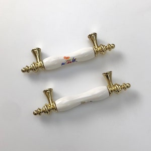May include: Two white ceramic cabinet knobs with gold accents and floral designs. The knobs have a decorative, ornate design.
