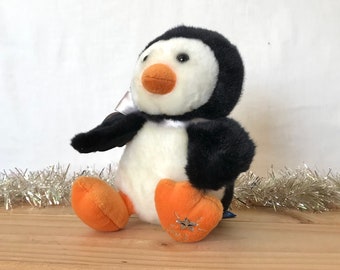 May include: A plush penguin toy with black and white fur, an orange beak, and orange feet. The penguin features a silver ribbon tied around its neck and a silver embroidered star design on the sole of its right foot, showcasing soft textures.