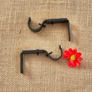 May include: Two black metal curtain rod brackets with a single screw on each bracket. The brackets are on a brown burlap background with a red flower.