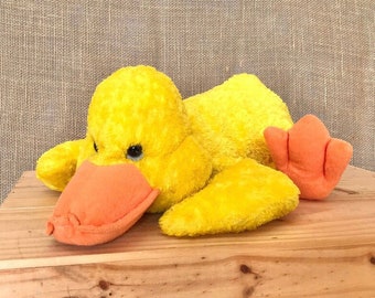 May include: A yellow plush duck toy with orange beak and feet. The duck is lying on its side on a wooden surface.