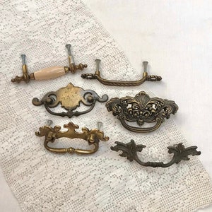 May include: Six antique cabinet knobs with ornate designs. The knobs are made of brass and have a variety of finishes, including polished brass, brushed brass, and antique brass. The knobs are in good condition and would be a great addition to any home.