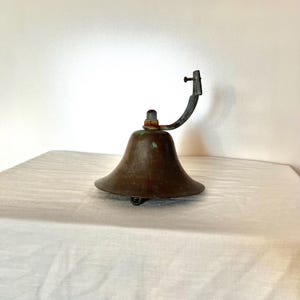 May include: A vintage copper bell with a metal bracket. The bell has a green patina and is mounted on a white surface.