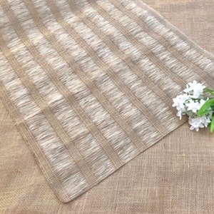 May include: A rectangular, beige woven placemat with vertical fringed stripes. The placemat is made of a natural fiber material. A sprig of white flowers with green leaves is in the corner.