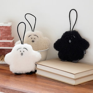 Puede incluir: Tres llaveros de peluche con forma de nube. Los llaveros son blancos, beige y negros. Los llaveros están colgados de una superficie de madera.