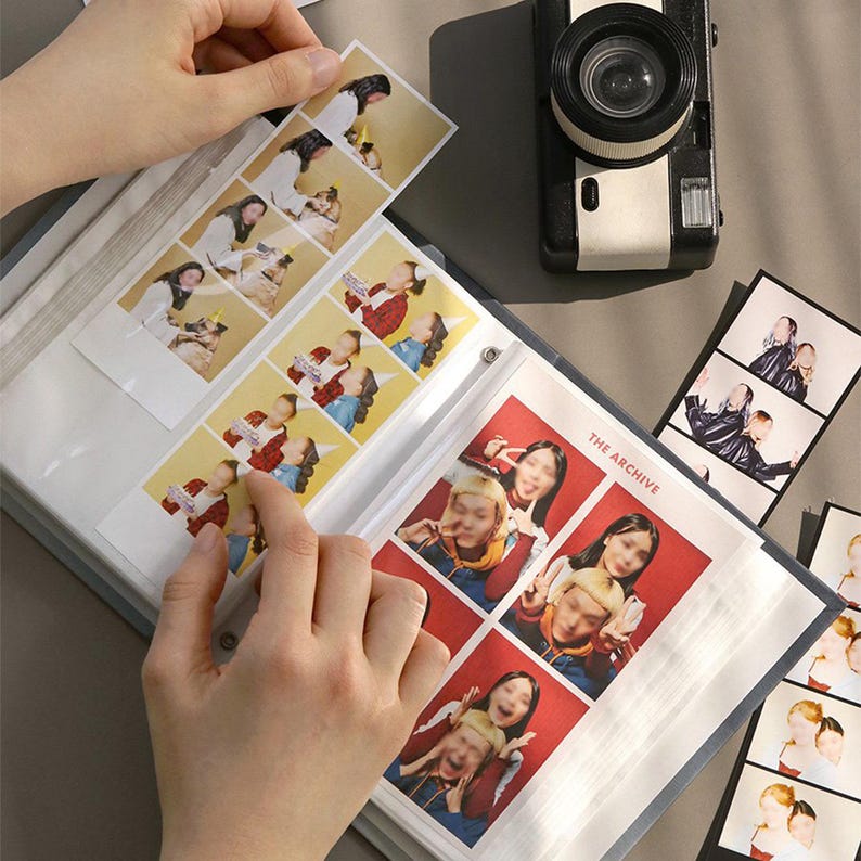 Op de afbeelding: Een zwart fotoalbum met doorzichtige plastic hoesjes om foto's in te bewaren. Het album heeft de tekst "The Archive" op de voorzijde. Het album is open om foto's van mensen te laten zien.