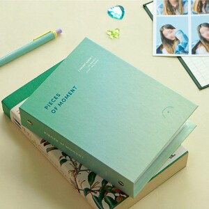 Op de afbeelding: Een groen en blauw fotoalbum met een ringband. Het album is gesloten en heeft de tekst "Pieces of Moment" op de voorzijde. Het album ligt op een boek met een bloemenpatroon.