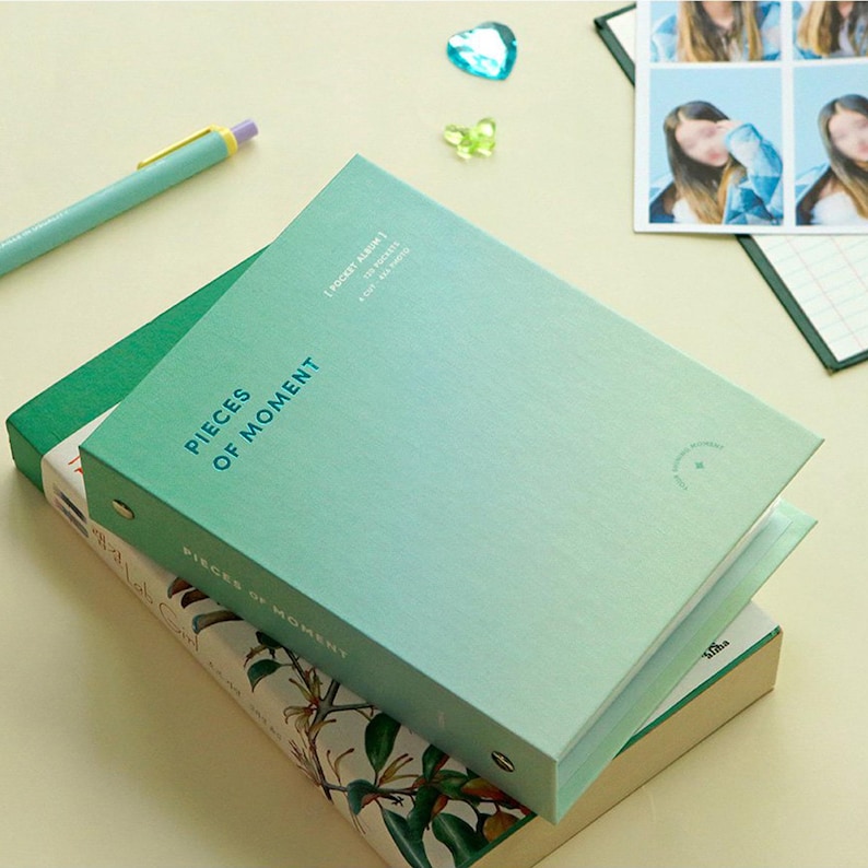 Op de afbeelding: Een groen en blauw fotoalbum met een ringband. Het album is gesloten en heeft de tekst "Pieces of Moment" op de voorzijde. Het album ligt op een boek met een bloemenpatroon.