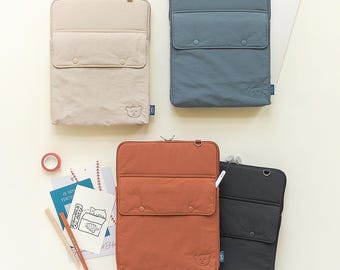 May include: Four laptop sleeves in different colours: beige, blue, orange, and black. Each sleeve has a front pocket and a small embroidered bear design. The sleeves are made of a soft, textured fabric.