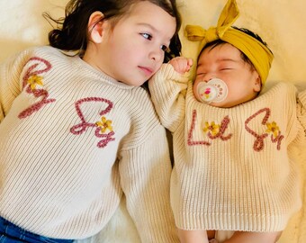 May include: Two sisters wearing matching cream knitted sweaters with embroidered flowers and the words "Big Sis" and "Lil Sis". The older sister is wearing blue jeans and has her hair in two buns. The younger sister is wearing a yellow headband and a pacifier.