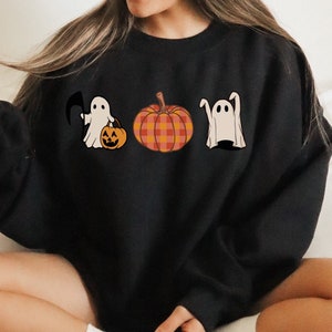 以下が含まれることがあります： 3体の幽霊とカボチャのグラフィックがプリントされた黒いスウェットシャツ。幽霊は白で黒い目。カボチャはオレンジで格子模様です