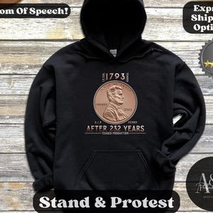 May include: Black hoodie with a front graphic featuring a copper-colored penny design. The design includes the text "1793", "IN GOD WE TRUST", "LIBERTY", "2025", and "AFTER 232 YEARS". The hoodie also has the text "Freedom Of Speech!" and "Stand & Protest".