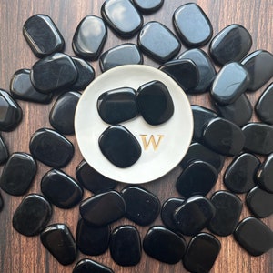 May include: A collection of smooth, black obsidian stones, some of which are arranged on a white plate with the letter "W" in gold lettering.