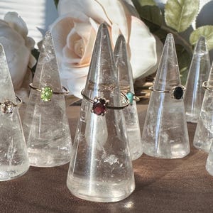 May include: A collection of rings with different colored gemstones displayed on clear crystal ring holders. The rings feature a variety of gemstones, including green, red, black, and turquoise.