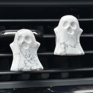 May include: Two white ghost-shaped car air fresheners. The ghosts have a marble-like texture with gray veining. Each ghost has a simple face with eye and mouth cutouts. They are positioned on a car vent.
