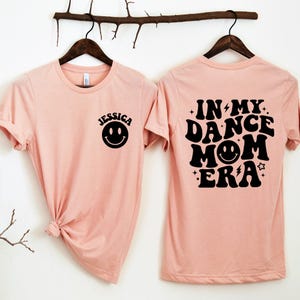Op de afbeelding: Een roze T-shirt met een zwart smiley-gezicht op de voorkant en de tekst "In My Dance Mom Era" met een zwart smiley-gezicht op de achterkant.